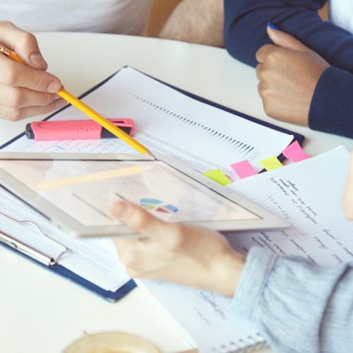 Working in team: young businesspeople discussing business plans and ideas, analyzing graphics and diagrams on paper. Man holding pen, pointing at tablet pc screen, explaining project strategy